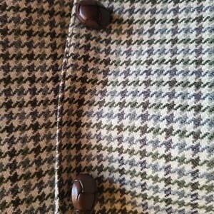 May include: Close-up of a tweed fabric with a houndstooth pattern in shades of green, gray, and beige. Two dark brown, leather-like buttons are visible on the left side, suggesting a coat or jacket.