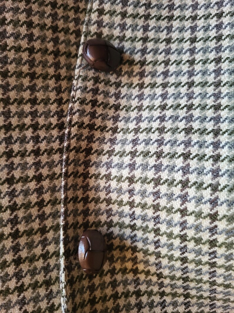 May include: Close-up of a tweed fabric with a houndstooth pattern in shades of green, gray, and beige. Two dark brown, leather-like buttons are visible on the left side, suggesting a coat or jacket.