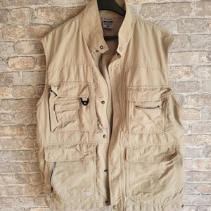 Könnte beinhalten: Eine beige Utility-Weste mit mehreren Taschen und einem Reißverschluss. Die Weste hat ein Columbia-Logo auf der linken Brust.