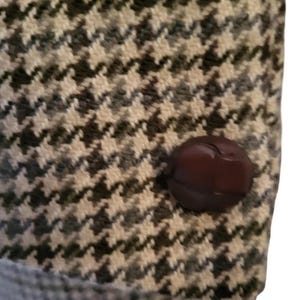 May include: Close-up of a houndstooth pattern fabric with a dark brown button. The fabric is a mix of black, white, and gray, creating a classic, textured design. The button is round and has a glossy finish.