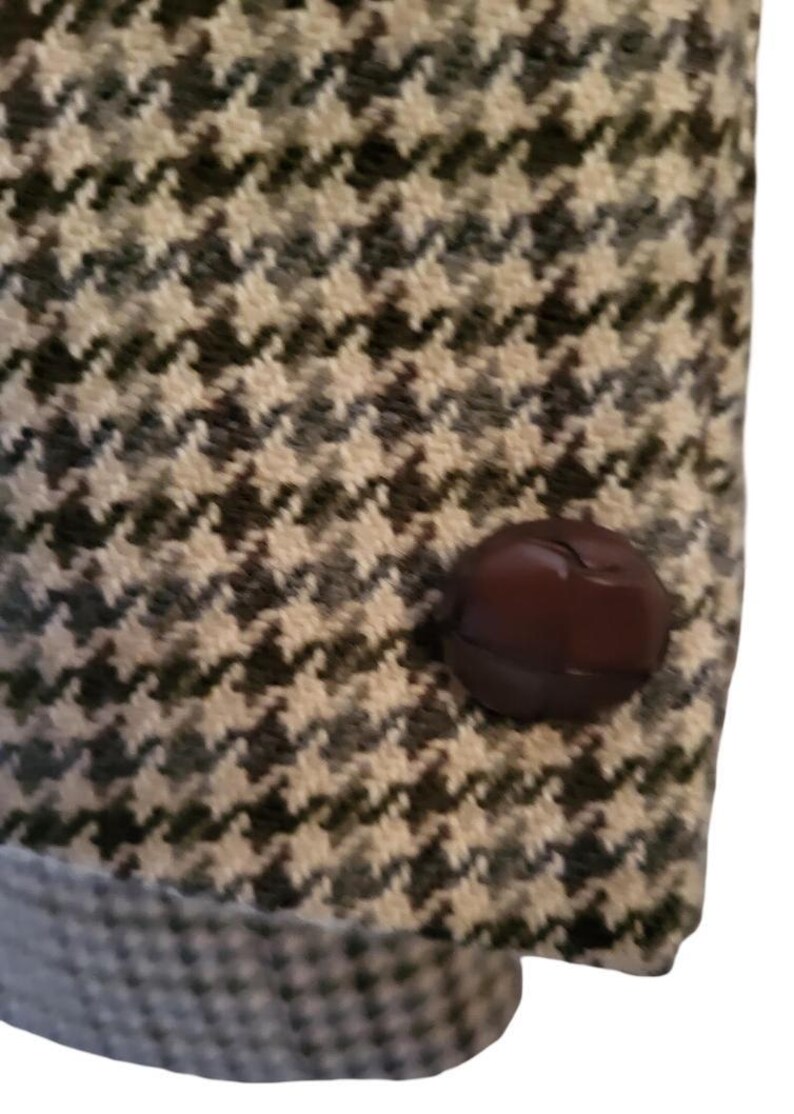 May include: Close-up of a houndstooth pattern fabric with a dark brown button. The fabric is a mix of black, white, and gray, creating a classic, textured design. The button is round and has a glossy finish.