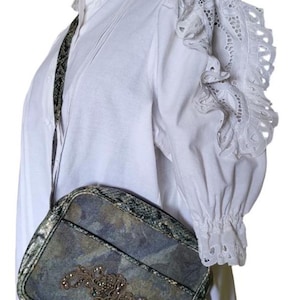 Könnte beinhalten: Eine Umhängetasche mit einem Tarnmuster in Blau-, Grün- und Grautönen. Die Tasche hat ein dekoratives Blumendesign mit Strasssteinen. Der Riemen hat ein dunkelgrünes Schlangenmuster. Die Tasche wird mit einer weißen Bluse mit Spitzendetails kombiniert.