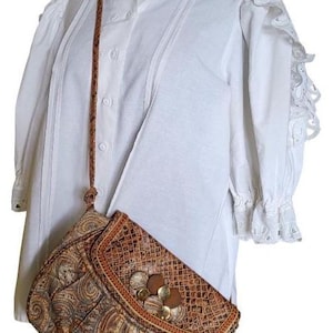 May include: A white blouse with a high ruffled collar and lace trim on the sleeves. A paisley-patterned handbag with a brown leather strap is worn over the shoulder. The bag features a decorative clasp.