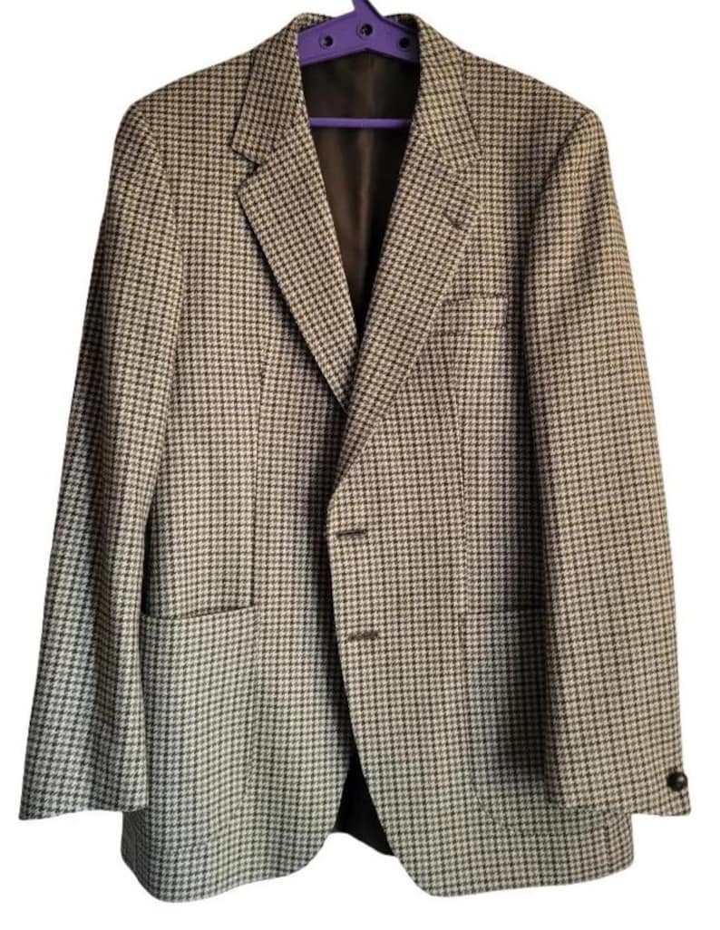 May include: A houndstooth patterned blazer in shades of brown and white, hanging on a purple hanger. The jacket features a notched lapel, two front pockets, and a two-button closure. The interior lining is a dark brown color.