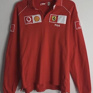 Könnte beinhalten: Rotes Langarm-Poloshirt mit weißen Akzenten und Kragen. Mit Sponsorenlogos wie Vodafone und Shell sowie einem Ferrari-Emblem. Das Shirt hat das Fila-Logo und deutet auf ein Sport- oder Rennsportthema hin.
