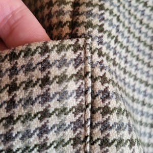 May include: Close-up of a houndstooth fabric in shades of green, brown, and cream. The fabric has a textured appearance with a diagonal pattern. The stitching is visible along the seam.