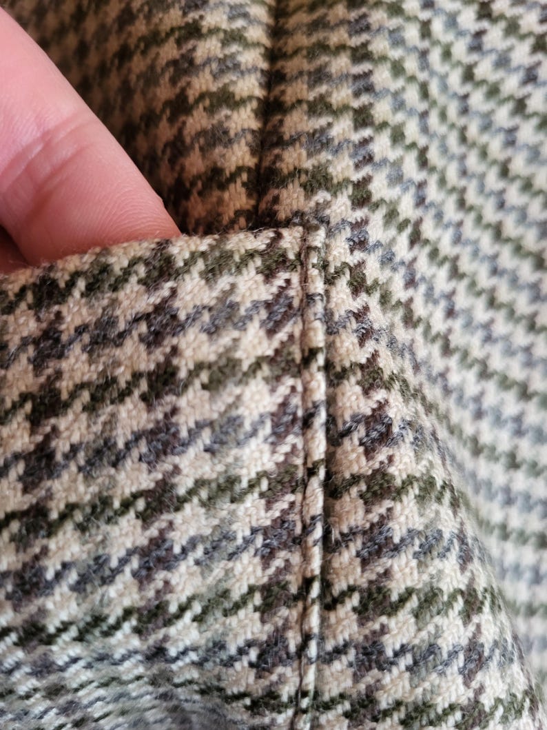 May include: Close-up of a houndstooth fabric in shades of green, brown, and cream. The fabric has a textured appearance with a diagonal pattern. The stitching is visible along the seam.