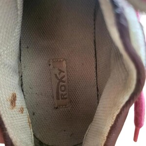 May include: Interior view of a well-worn sneaker. The shoe's interior features a light brown insole with the brand name "ROXY" visible. The shoe's lining is a light beige color, and the laces are pink.