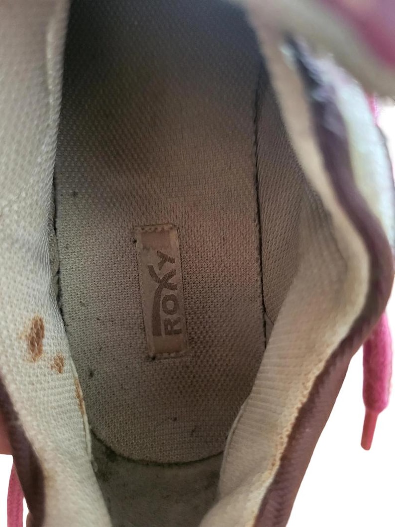 May include: Interior view of a well-worn sneaker. The shoe's interior features a light brown insole with the brand name "ROXY" visible. The shoe's lining is a light beige color, and the laces are pink.
