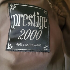 May include: A close-up of a black label with the words "prestige 2000" in silver lettering. The label also states "100% Lambswool". The label is sewn onto a brown fabric.