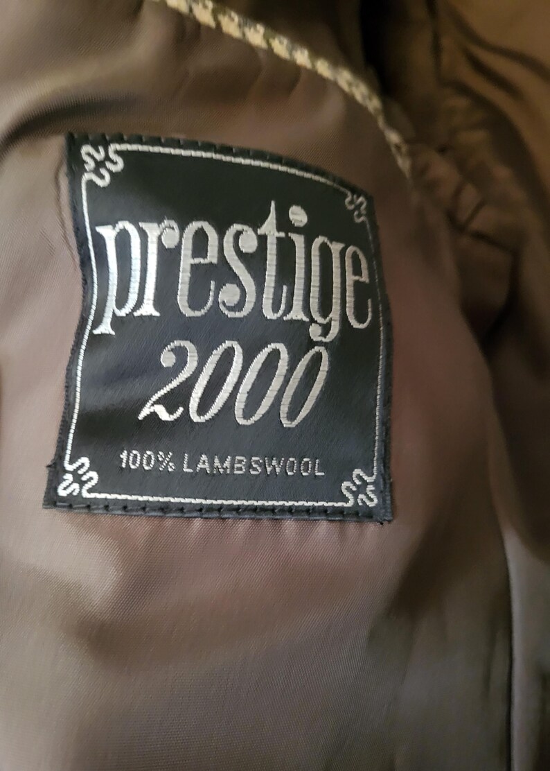 May include: A close-up of a black label with the words "prestige 2000" in silver lettering. The label also states "100% Lambswool". The label is sewn onto a brown fabric.
