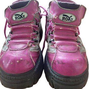 May include: Pair of well-worn, pink and gray Roxy high-top boots. The boots have a chunky black sole, pink laces, and the Roxy logo on the side. The leather shows signs of wear and tear.