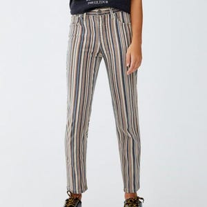 May include: Straight-leg jeans with vertical stripes in beige, brown, and navy blue. The jeans feature a classic five-pocket design. The model is wearing black platform shoes. The top is a black t-shirt with the text "BACK IN BLACK 1980 US TOUR".