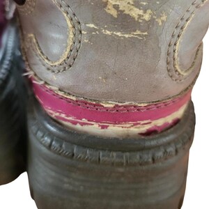 May include: Close-up of a worn brown boot with a thick, dark sole. The boot's upper shows signs of wear and tear, with peeling paint revealing a pink layer beneath. The stitching is visible along the edges.