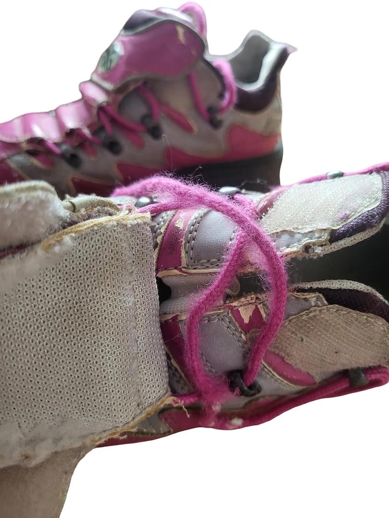 May include: Close-up of a pair of worn athletic shoes. The shoes are primarily gray and pink, with pink laces and accents. The shoes have a hook-and-loop closure and show signs of wear and tear.