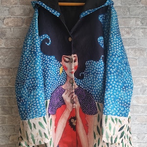 Puede incluir: Un abrigo con capucha con una ilustración artística de una mujer con cabello negro y una flor roja. El abrigo tiene un diseño azul y blanco en las mangas y una capucha negra. El abrigo está abotonado.