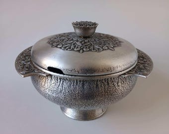May include: Silver-colored, ornate serving dish with a lid. The dish has a textured surface and decorative handles. The lid features a floral design and a small, flower-shaped knob. A small opening is visible on the side of the dish.