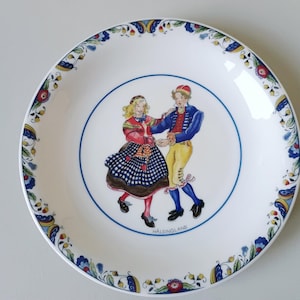 Puede incluir: Un plato de cerámica blanca con una ilustración central de dos figuras bailando con ropa tradicional. El borde está decorado con un colorido patrón floral en azul, rojo y amarillo. La palabra HÄLSINGLAND está impresa en letras negras pequeñas debajo de las figuras.