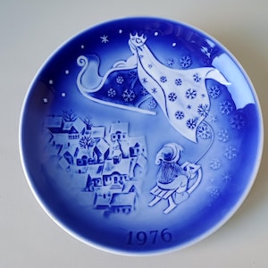 May include: A blue and white Christmas plate with a winter scene. A woman in a white robe is flying in a sleigh pulled by reindeer over a small town. The plate has the year 1976 printed on the bottom.