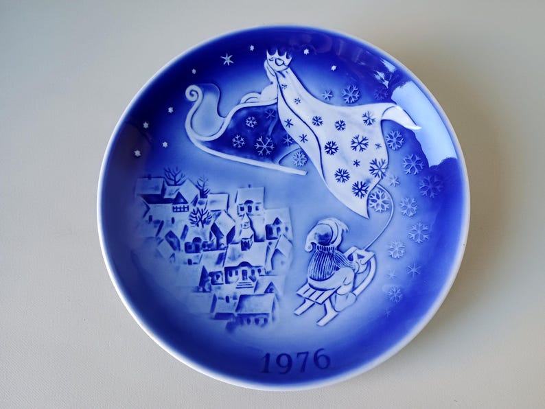 May include: A blue and white Christmas plate with a winter scene. A woman in a white robe is flying in a sleigh pulled by reindeer over a small town. The plate has the year 1976 printed on the bottom.
