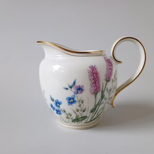 May include: White porcelain creamer with a gold rim and handle. The creamer is decorated with a floral design featuring blue and purple flowers and pink lavender-like blooms. A classic piece for serving cream or milk.