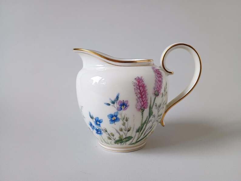 May include: White porcelain creamer with a gold rim and handle. The creamer is decorated with a floral design featuring blue and purple flowers and pink lavender-like blooms. A classic piece for serving cream or milk.