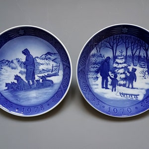 Puede incluir: Dos platos decorativos azules y blancos. Uno representa a una persona con perros de trineo en un paisaje nevado, el otro a una familia con un trineo en un bosque invernal. Ambos platos tienen el a&ntilde;o impreso.
