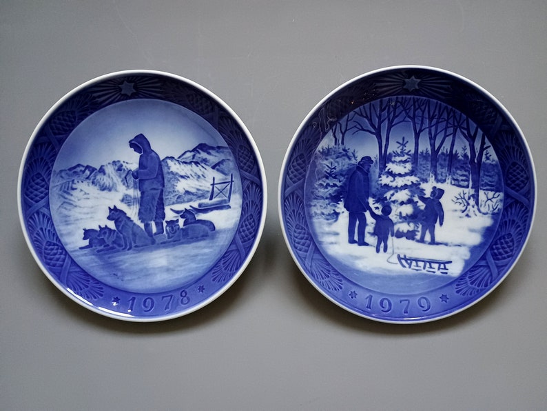 Puede incluir: Dos platos decorativos azules y blancos. Uno representa a una persona con perros de trineo en un paisaje nevado, el otro a una familia con un trineo en un bosque invernal. Ambos platos tienen el a&ntilde;o impreso.