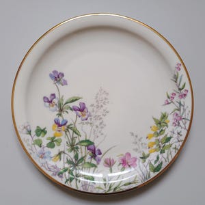 May include: A white ceramic plate with a gold rim, decorated with a floral pattern of purple, yellow, and pink flowers. The flowers are arranged around the edge of the plate, creating a delicate and elegant design.