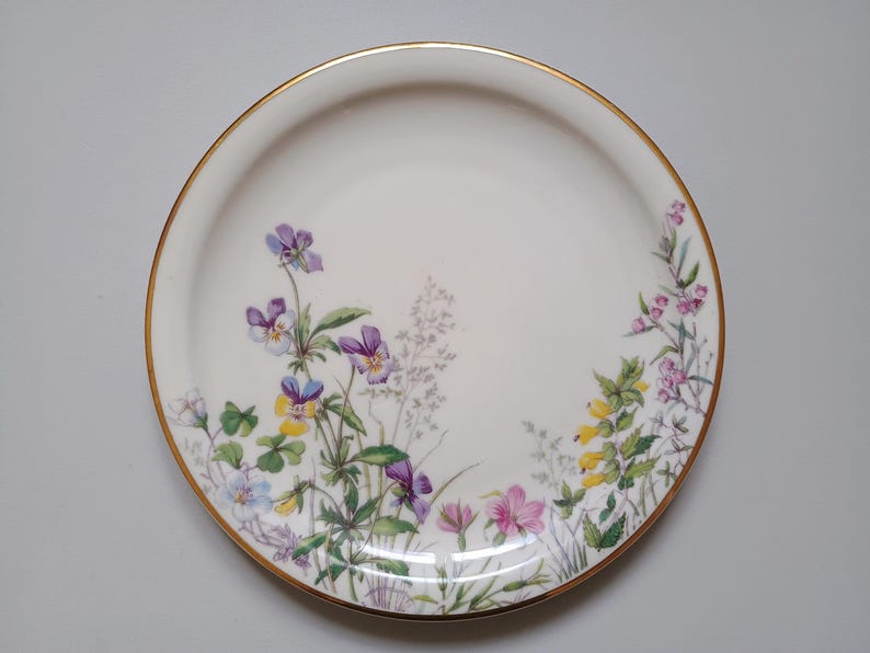 May include: A white ceramic plate with a gold rim, decorated with a floral pattern of purple, yellow, and pink flowers. The flowers are arranged around the edge of the plate, creating a delicate and elegant design.