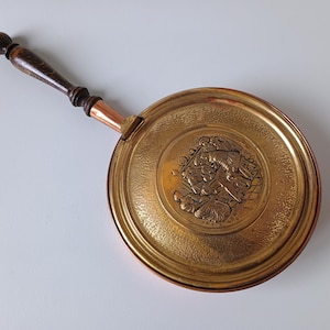 Pode incluir: Um aquecedor de cama redondo de latão com um cabo longo de madeira castanho-escura e um pescoço de cobre. A tampa circular de latão apresenta uma cena central em relevo com figuras em trajes históricos, rodeada por uma superfície de metal martelado.