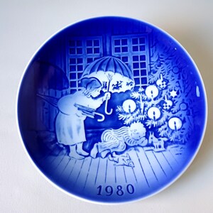 May include: Blue and white Christmas plate with a scene of a child holding an umbrella and looking at a Christmas tree with lit candles. The plate has the year 1980 printed on the bottom.