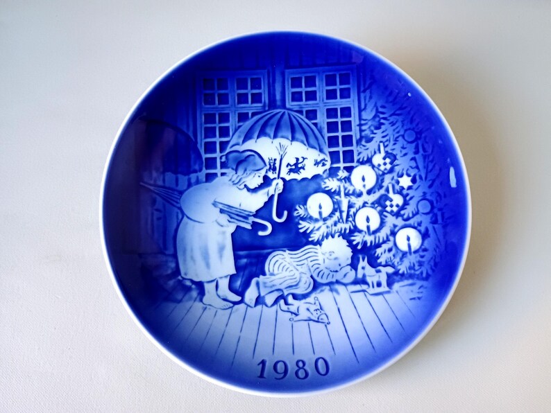 May include: Blue and white Christmas plate with a scene of a child holding an umbrella and looking at a Christmas tree with lit candles. The plate has the year 1980 printed on the bottom.