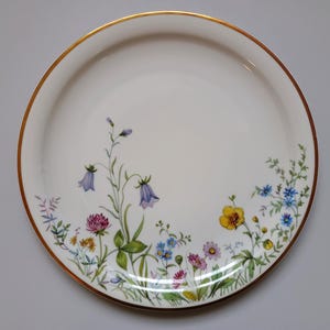 May include: A white ceramic plate with a gold rim, decorated with a floral design. The flowers include purple bellflowers, yellow blooms, and pink clover, with green leaves and stems. The plate is round.
