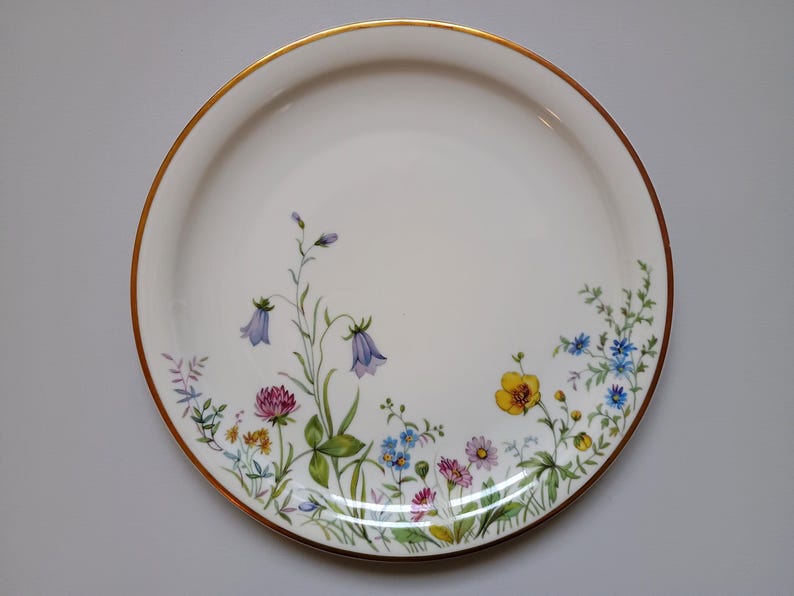May include: A white ceramic plate with a gold rim, decorated with a floral design. The flowers include purple bellflowers, yellow blooms, and pink clover, with green leaves and stems. The plate is round.