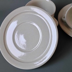 K&ouml;nnte beinhalten: Eine wei&szlig;e Untertasse aus Keramik mit einem erh&ouml;hten Rand und einer glatten, gl&auml;nzenden Oberfl&auml;che. Die Untertasse hat ein konzentrisches Kreisdesign.