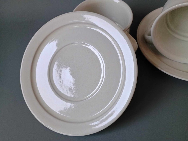 K&ouml;nnte beinhalten: Eine wei&szlig;e Untertasse aus Keramik mit einem erh&ouml;hten Rand und einer glatten, gl&auml;nzenden Oberfl&auml;che. Die Untertasse hat ein konzentrisches Kreisdesign.