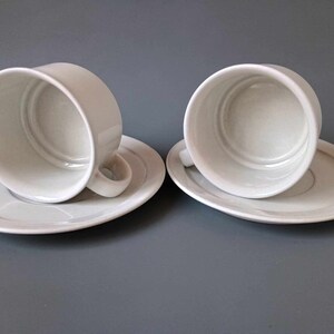 K&ouml;nnte beinhalten: Zwei wei&szlig;e Keramik-Kaffeetassen mit Untertassen. Die Tassen sind leer und die Untertassen sind schlicht wei&szlig;.