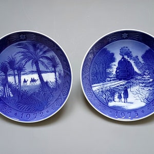 Puede incluir: Dos platos decorativos azules y blancos. Uno representa una escena des&eacute;rtica con palmeras y camellos, el otro un tren en un paisaje nevado. Ambos platos tienen un dise&ntilde;o de estrella y los a&ntilde;os 1972 y 1973.