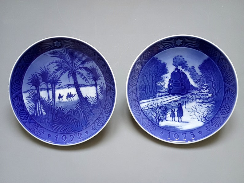Puede incluir: Dos platos decorativos azules y blancos. Uno representa una escena des&eacute;rtica con palmeras y camellos, el otro un tren en un paisaje nevado. Ambos platos tienen un dise&ntilde;o de estrella y los a&ntilde;os 1972 y 1973.