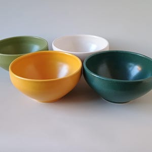 May include: Four ceramic bowls in shades of green, yellow, white, and dark green. The bowls are round with a smooth, glossy finish. The bowls are empty and arranged on a light gray surface.