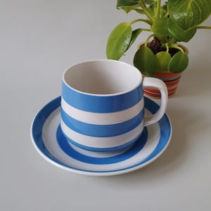 Pode incluir: Um conjunto de chávena e pires em cerâmica com um padrão de riscas horizontais em azul brilhante e branco. A chávena tem uma pega lateral resistente e o pires apresenta anéis concêntricos a condizer em azul e branco, criando um design coeso.