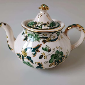 May include: A cream-coloured ceramic teapot featuring a detailed floral pattern in shades of dark green, light green, and gold. The teapot has a matching lid with a gold-trimmed finial, a curved handle, and a spout, all accented with gold.