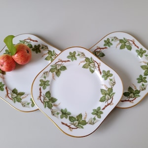 Può includere: Tre piatti quadrati in porcellana bianca con angoli arrotondati e bordi dorati. Ogni piatto presenta un motivo botanico di foglie verdi, steli marroni e piccoli fiori bianchi. Due piccole mele rosse con una foglia verde poggiano su un piatto.
