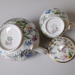 May include: A white porcelain tea set with a floral pattern featuring blue, purple, and yellow flowers. The set includes a teapot, a teacup, and a lid with a gold trim. The teapot has a curved spout and handle.
