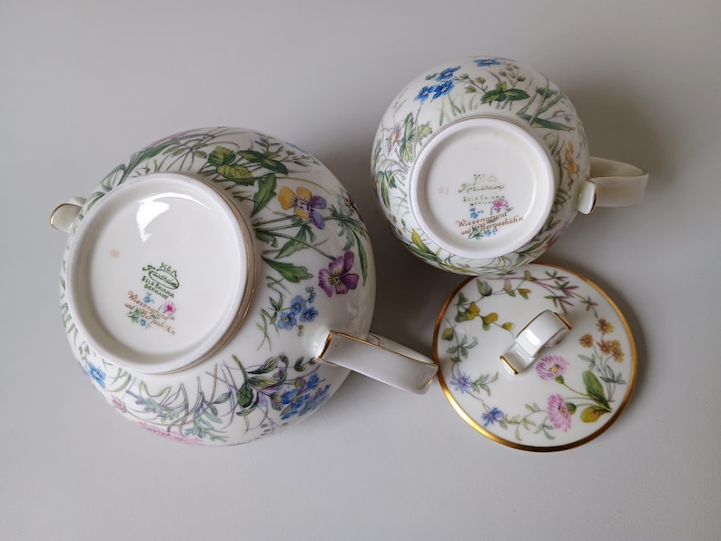 May include: A white porcelain tea set with a floral pattern featuring blue, purple, and yellow flowers. The set includes a teapot, a teacup, and a lid with a gold trim. The teapot has a curved spout and handle.