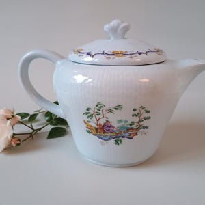 May include: A white porcelain teapot with a ribbed texture, featuring a colourful hand-painted scene of a figure in a landscape on the body and floral accents on the lid. The teapot has a curved handle, a spout, and a matching lid with a finial.