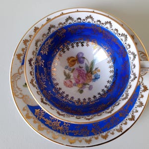 Può includere: Un set di tazza da tè e piattino in porcellana con un bordo blu cobalto vibrante e dettagli in filigrana dorata. Il centro della tazza e il piattino mostrano un delicato bouquet di fiori multicolori su uno sfondo bianco.
