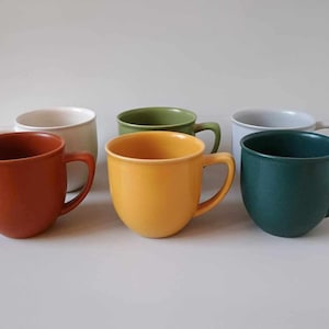Può includere: Sei tazze in ceramica di diversi colori: bianco, verde, giallo, arancione e due tonalità di verde. Le tazze hanno una forma arrotondata e un manico sul lato.