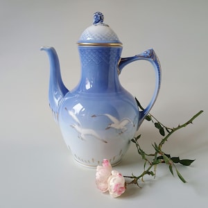 Puede incluir: Una tetera de porcelana en color azul claro y blanco con una escena pintada de gaviotas en vuelo. La tetera tiene una tapa texturizada con un pequeño remate, un asa curva y un pico largo. Detalles dorados adornan la tapa, el asa y la base.